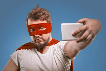 超级英雄男人。需要自拍移动手机自信男人。超级英雄服装红色的面具斗篷提出了褪了色的牛仔布蓝色的背景使自拍照片智能手机