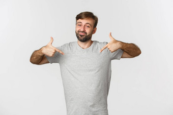 肖像英俊的有胡子的男人。灰色的t恤指出标志微笑推荐品牌站白色背景