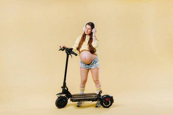 怀孕了女孩黄色的衣服耳机电踏板车孤立的黄色的背景