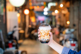 年轻的女孩持有显示杯棕色（的）糖味木薯珍珠泡沫牛奶茶晚上市场台湾背景关闭散景