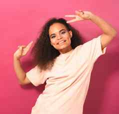 年轻的非洲美国女孩非洲式发型头发跳舞手势和平手解除积极相机穿桃色的t恤孤立的粉红色的背景美概念广场镜头