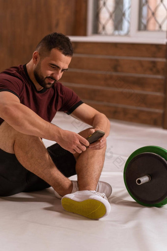英俊的男人。坐着地板上智能手机手黑色的绿色语气健身杠铃设备重量培训概念体育设备培训健康的生活方式概念