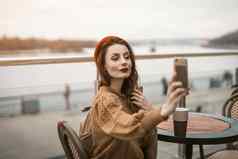 美丽的年轻的女人需要自拍智能手机杯咖啡法国女人穿红色的贝雷帽坐着阳台餐厅咖啡馆背景秋天城市城市