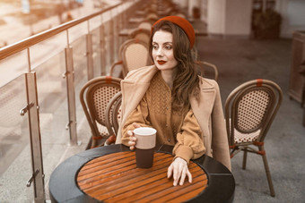 深思熟虑的记忆年轻的巴黎女人坐着在户外餐厅阳台持有咖啡杯子肖像时尚的年轻的女人穿秋天外套红色的贝雷帽在户外