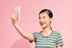 自拍！快乐的年轻的亚洲女人持有移动电话使自拍聪明的电话站粉红色的背景