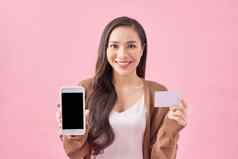 肖像微笑年轻的女人持有空白屏幕移动电话显示信贷卡孤立的粉红色的背景