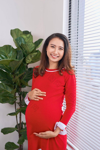美丽的怀孕了妈妈。红色的衣服摆姿势装饰工作室微笑相机妈妈站窗口