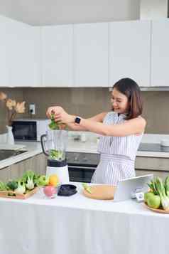 健康的亚洲女人享受使绿色蔬菜排毒净化绿色水果奶昔搅拌机厨房首页节食概念健康的生活方式