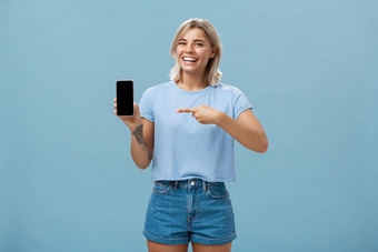 非常滑稽的照片娱乐有吸引力的快乐女人公平头发休闲t恤牛仔布短裤显示智能手机相机指出设备屏幕微笑广泛的蓝色的墙
