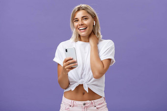 害羞的有吸引力的时尚的善于交际女人白色t恤触碰脖子胆小尴尬的微笑广泛的穿无线耳机听歌曲智能手机紫色的墙