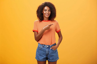 显示完美的的地方广告迷人的无忧无虑的自信年轻的时尚的非裔美国人女人非洲式发型发型持有手口袋里随便指出橙色墙