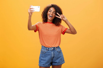 女孩使视频视频博客品牌智能<strong>手机发布</strong>互联网著名的站橙色背景摆姿势自拍盯着小工具屏幕显示和平胜利手势