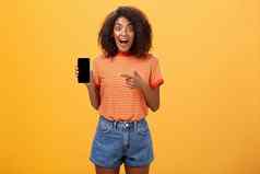 深刻的印象着迷时尚的苗条的非洲美国卷发女人条纹t恤下降下巴惊讶的是持有太棒了智能手机指出设备屏幕橙色墙