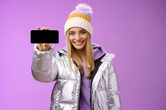 自信友好的好看的金发碧眼的女孩户外银闪闪发光的夹克他持有智能手机水平显示移动电话显示自信的微笑推荐应用程序紫色的背景