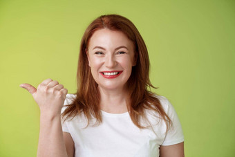 访问商店快乐的愉快的友好的迷人的红色头发的人中年女人企业家邀请退房促销微笑幸福的真诚的种类露齿而笑指出左拇指站绿色背景