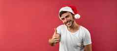 年轻的英俊的高加索人的家伙白色t恤圣诞老人帽子站红色的背景工作室显示拇指