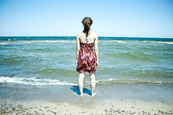 女人长棕色（的）头发穿红色的背心裙站没膝的水海滩波腿上脚踝