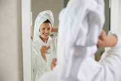 快乐微笑女孩喝持有玻璃水站前面镜子浴室女孩穿袍采取淋浴概念健康的生活方式