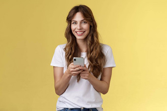热情的迷人的善于交际年轻的女孩消息传递朋友发送照片社会媒体持有智能手机相机幸福的友好的微笑站黄色的背景休闲装