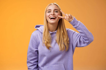 有吸引力的友好的无忧无虑的千禧金发碧眼的女孩紫色的连帽衫有趣的朋友笑快乐显示和平胜利嬉皮标志喜欢完美的寒冷的春天天气橙色背景