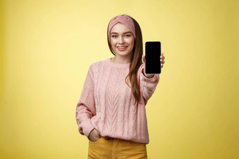 智能手机迷人的即将离任的年轻的漂亮的女人针织毛衣头巾扩展手电话显示小工具屏幕微笑推荐手机黄色的墙