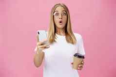 女孩接收太棒了消息智能手机喝咖啡杯咖啡馆分享新闻朋友折叠嘴唇哇声音盯着惊讶相机持有手机粉红色的墙