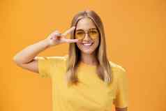 可爱的乐观友好的年轻的高加索人女人公平头发黄色的t恤太阳镜显示和平标志眼睛微笑高高兴兴地相机采取拍摄橙色背景