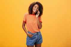 时尚的皮肤黝黑的女孩使休闲电话调用朋友告诉细节浪漫的日期站很高兴无忧无虑的橙色背景条纹t恤盯着左露齿而笑