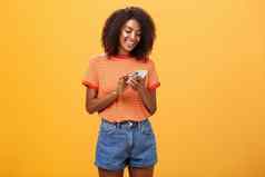 时尚的无忧无虑的女孩发短信朋友站很高兴橙色墙时尚的牛仔布短裤打字消息滚动新闻互联网智能手机盯着设备屏幕微笑