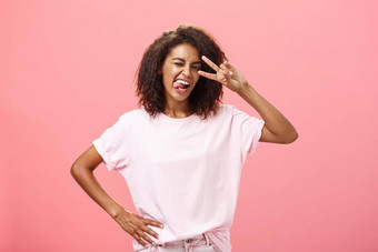 害怕表达快乐的有魅力的非洲美国女人t恤非洲式发型发型显示舌头开玩笑地大胆的使和平标志眼睛眨眼摆姿势粉红色的背景