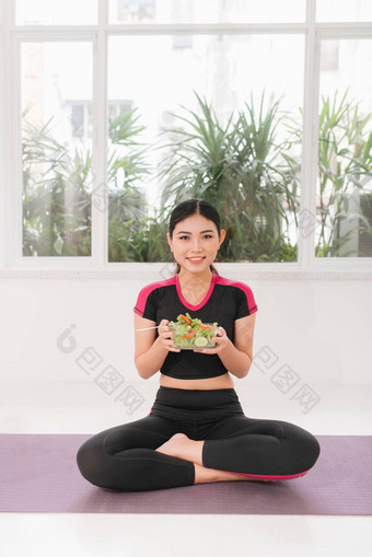 年轻的亚洲女人健身穿素食者健康的沙拉坐着地板上白色墙