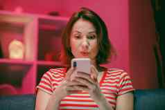 快乐年轻的女人持有聪明的电话手机屏幕笑享受移动应用程序购物有趣的玩游戏聊天社会媒体坐沙发上首页