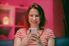 年轻的女人持有聪明的电话手机屏幕笑享受移动应用程序购物有趣的玩游戏聊天社会媒体坐沙发上首页