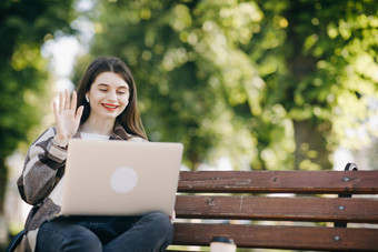 女孩挥舞着手网络摄像头移动PC坐着公园板凳上学生网络摄像头移动PC视频闲谈，聊天朋友高加索人女人微笑坐着板凳上午餐时间在户外