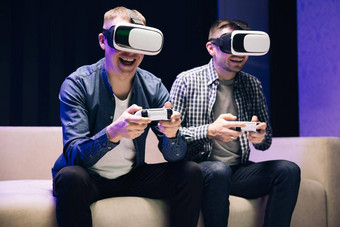兴奋有趣的年轻的游戏玩家穿虚拟现实眼镜持有控制器玩视频游戏喜出望外但朋友赢得电子游戏有趣的沙发庆祝胜利首页