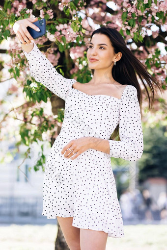 女显示积极的脸情绪年轻的女人夏天白色衣服采取自拍肖像照片智能手机模型摆姿势公园樱花树背景