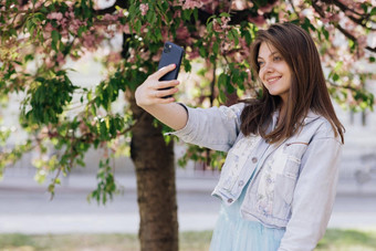 年轻的微笑女人采取自拍肖像照片智能手机模型摆姿势公园樱花树背景女显示积极的脸情绪