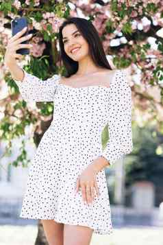 年轻的微笑女人夏天白色衣服采取自拍肖像照片智能手机模型摆姿势公园樱花树背景女显示积极的脸情绪