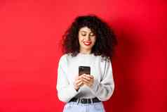 微笑欧洲女人阅读移动电话屏幕发短信消息智能手机站运动衫红色的背景