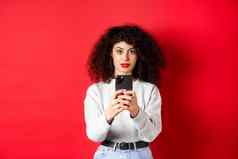 年轻的女人卷曲的头发记录视频智能手机采取照片移动电话相机站红色的背景