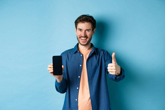 英俊的年轻的男人。显示拇指手势空智能手机屏幕推荐应用程序公司站蓝色的背景