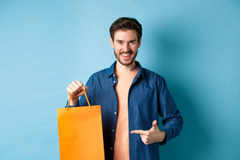 微笑高加索人男人。指出手指橙色购物袋买了项目站蓝色的背景
