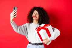 快乐年轻的女人采取自拍情人节一天礼物持有现在拍摄智能手机摆姿势红色的背景