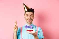 假期庆祝活动概念快乐年轻的男人。享受生日聚会，派对穿锥他交叉手指使bday蛋糕蜡烛粉红色的背景