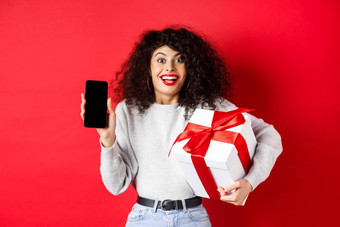 情人节情人一天兴奋微笑女人卷曲的黑暗头发显示智能手机空屏幕持有惊喜礼物假期显示在线促销红色的背景