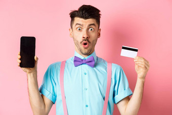 电子商务购物概念惊讶男人。显示空白智能<strong>手机屏幕</strong>塑料信贷卡站惊讶粉红色的背景