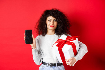 美丽的女人卷曲的头发显示购物<strong>应用</strong>程序空智能手机屏幕持有礼物<strong>包装</strong>节日盒子站红色的背景