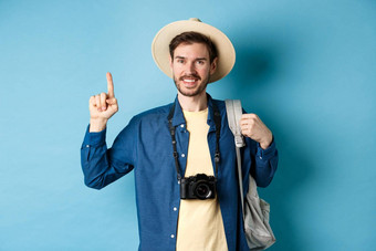 英俊的快乐旅游夏天他持有背包相机指出手指标志推荐旅行机构的地方假期蓝色的背景