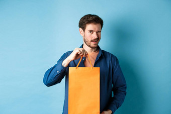 快乐年轻的男人。显示橙色购物袋眨眼推荐商店折扣站蓝色的背景
