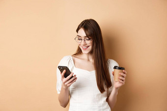 快乐女孩<strong>聊天</strong>电话喝咖啡穿眼镜白色上衣微笑智能手机屏幕米色背景